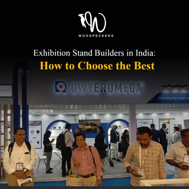 Exhibition booth fabricated for DwyerOmega by Woodpeckers in India, featuring illuminated 3D channel letters, arched wall display alcoves, track spotlights, and active trade show attendees.